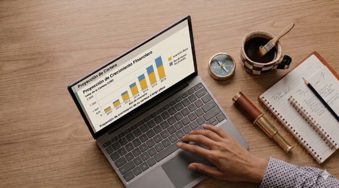 Ilustración desde arriba de una persona escribiendo en una laptop en un escritorio de madera. La pantalla muestra un gráfico de crecimiento financiero titulado "Provisión de Crecimiento Financiero" con años de 2010 a 2018. Alrededor de la laptop hay herramientas de análisis y exploración: una brújula, un telescopio de latón, un cuaderno abierto con notas y un lápiz, y una taza de café con un pincel dentro.